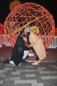 Jason kneels before Amberly amidst a glowing holiday light display shaped as an ornament. Their winter attire and tender embrace embody joy and commitment.